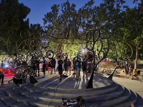 An image of the first stop at the SJBP. People are standing on a platform with circular sculptures above them. They are holding their bicycles up in the air. More people with bicycles are in the background. 